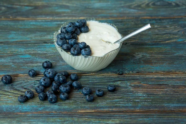 stock image Glass bowl with yogurt, blueberries and a teaspoon on a wooden blue aged background