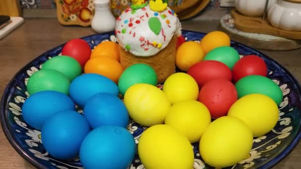 五颜六色的复活节彩蛋放在盘子里 盘子里放着复活节蛋糕 上面装饰着一只黄色的鸡 背景是切菜板和其他厨房用具 — 图库视频影像