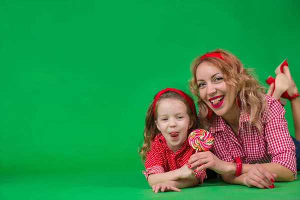 Mother and daughter dressed in pin-up style lie on a green background holding multi-colored caramel on a stick and show tongues