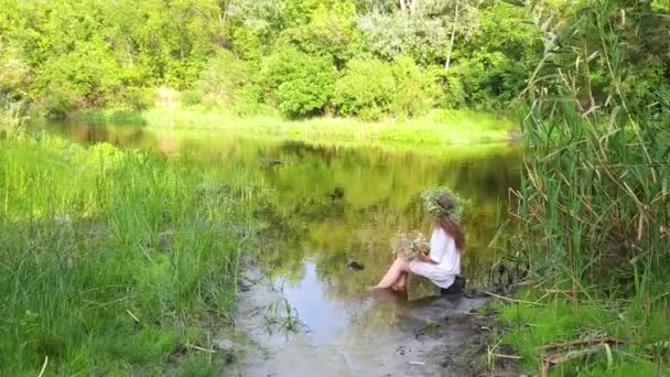 Dziewczyna Białej Koszuli Wieńcem Świeżych Ziół Siedzi Brzegu Rzeki Nogami — Wideo stockowe