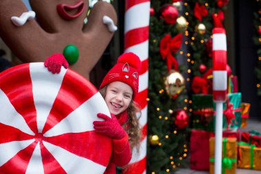 Kırmızılı bir kız çocuğu büyük bir karamel ve yeni yıl süslemelerinin önünde hediyelerle duruyor. Mutlu Noeller.