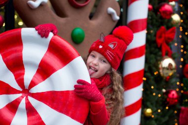 Kırmızı giyinmiş altı yaşında sarışın bir kız, Noel ağacının önünde çelenkle kocaman bir karamel yalıyor. Mutlu Noeller.