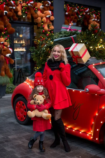 A mother with a six-year-old daughter stand near a red car in New Year's decorations with teddy bears. Merry Christmas