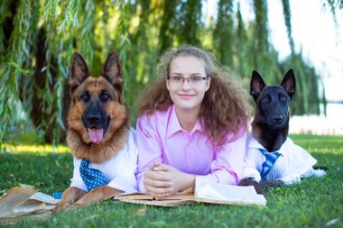 Alman çoban köpeği ve Belçikalı (Malinois) beyaz gömlekli, gözlüklü, pembe gömlekli, yeşil çimlerde oturan ve parkta kitap okuyan bir kızla.