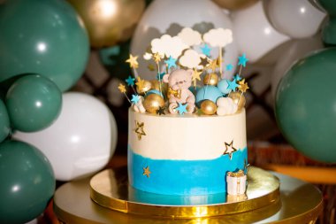 Küçük bir ayıyla dekore edilmiş mavi ve beyaz bir pasta çubuklardaki yıldızlar ve bulutlar ve şişme beyaz ve yeşil balonların arka planındaki mavi ve mavi balonlar.