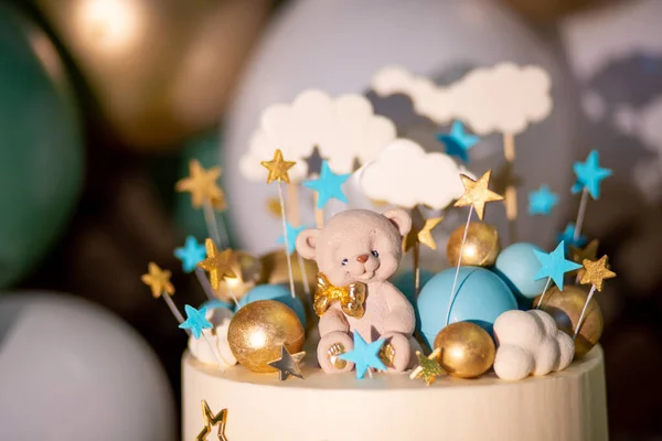 Blue and white cake decorated with a small bear, stars and clouds on sticks and gold and blue balloons on a background of inflatable white and green balloons