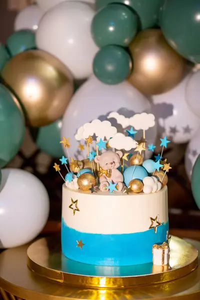 Blue and white cake on a gold stand decorated with a small bear, stars and clouds on sticks and gold and blue balloons on a background of inflatable white and green balloons