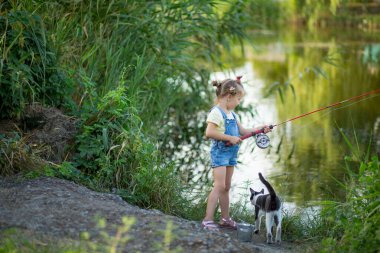 Üç yaşında kot şortlu sarışın bir kız nehirde siyah beyaz bir kediyle balık yakalar.