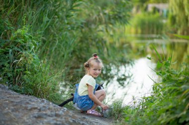 Üç yaşında kot şortlu sarışın bir kız nehirde siyah beyaz bir kediyle balık yakalar.