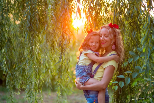 Blonde mother and three-year-old daughter in denim shorts with straps with red bows against the backdrop of hanging willow branches in the rays of the setting sun. Mother holds daughter in her arms