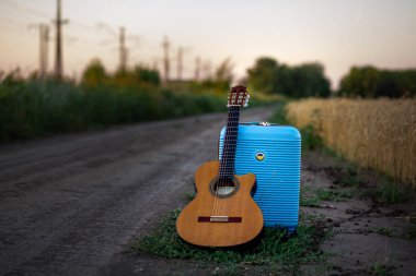 Üzerinde sarı-mavi bayrak bulunan büyük mavi bir bavulu olan gitar akşamları bir buğday tarlasının yanında toprak bir yolda duruyor.