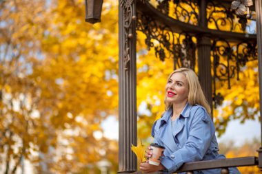 Gri yağmurluklu bir kız, elinde bir fincan kahve ve bir sonbahar parkında bir taraçada sarı bir akçaağaç yaprağı.