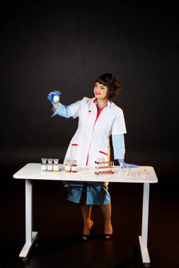 Siyah arka planda laboratuvar önlüklü bir öğretmen. Bir takım kimyasal bileşikler ve çeşitli cam test tüpleri var.