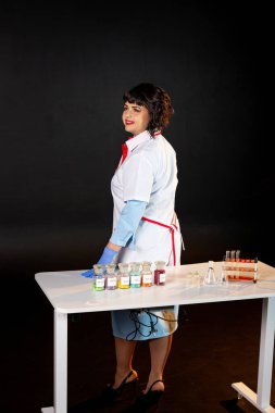 Siyah arka planda laboratuvar önlüklü bir öğretmen. Bir takım kimyasal bileşikler ve çeşitli cam test tüpleri var.