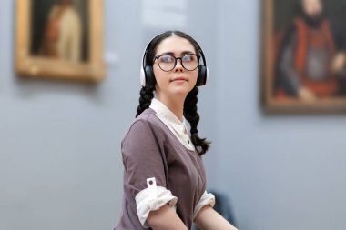 Portrait of young Caucasian pretty woman wearing headphones and contemplates ancient artefacts. Student visiting arts exhibition. Concept of culture education.