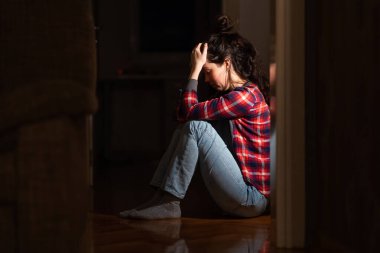 Panic disorder and obsessive-compulsive disorder. Suffering young Caucasian woman sits down on floor in despair in dark living room. Concept of psychology and mental problems.