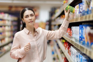 Visit grocery store. Portrait of young pretty caucasian woman takes food from upper shelve. Showcase at background. Concept of shopping and consumerism.
