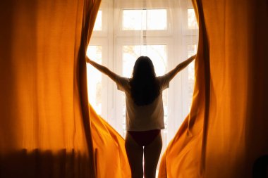 New day. Silhouette of woman opening orange curtains in morning at window. Back view. Concept of freedom, hope and happiness.