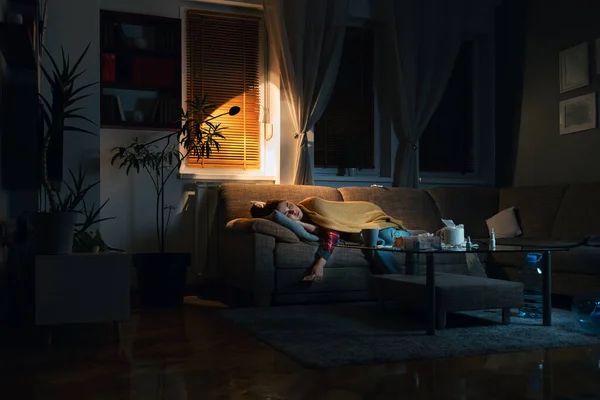 Depressed sleepy young Caucasian woman covered blanket lies on sofa in dark living room with electric light. Copy space. Concept of psychology illness and disease.
