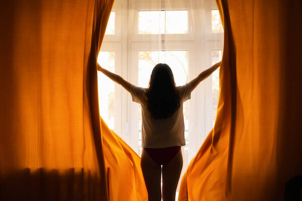 New day. Silhouette of woman opening orange curtains in morning at window. Back view. Concept of freedom, hope and happiness.