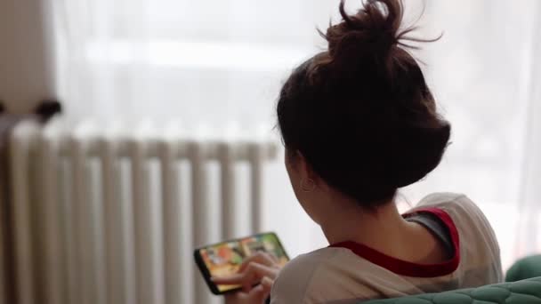若い女性はソファの上に家に座って 彼女のスマートフォンでオンラインゲームで遊んでいます バックビュー ソーシャルメディアやオンラインアプリの概念 — ストック動画