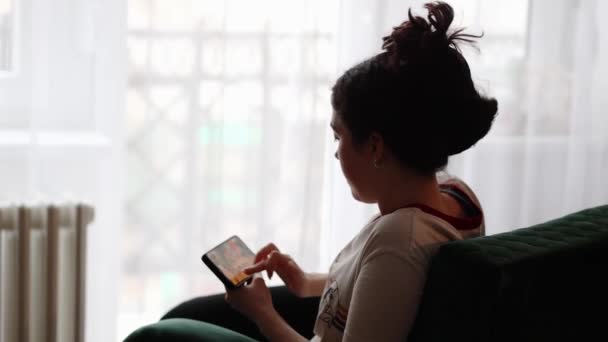 若い女性はソファの上に家に座って 彼女のスマートフォンでオンラインゲームで遊んでいます 後ろの景色 ソーシャルメディアやオンラインアプリの概念 — ストック動画