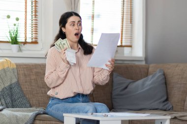 Purchase of real estate. Shocked young caucasian woman sitting on sofa, holds money and reading documents. Concept of leasing, business and mortgage.