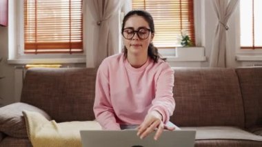 Freelance and remote work. Pretty young Caucasian woman wearing eyeglasses opens laptop. Zoom out. Home office with sofa and windows in background.