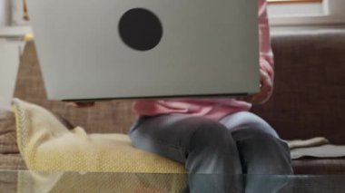 Freelance and remote work. Close up of smiling young Caucasian woman wearing eyeglasses take laptop and lean on sofa puts legs on table. Zoom out. Home office, working with comfort.