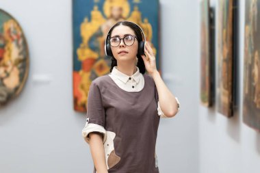 Portrait of young Caucasian pretty woman wearing headphones and contemplates ancient artefacts. Student visiting arts exhibition. Concept of education and culture.