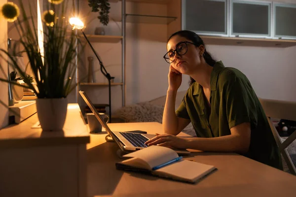 Online education. Portrait of boring young Caucasian woman student working at laptop. Evening in office with electric light. Concept of freelance and remote work.