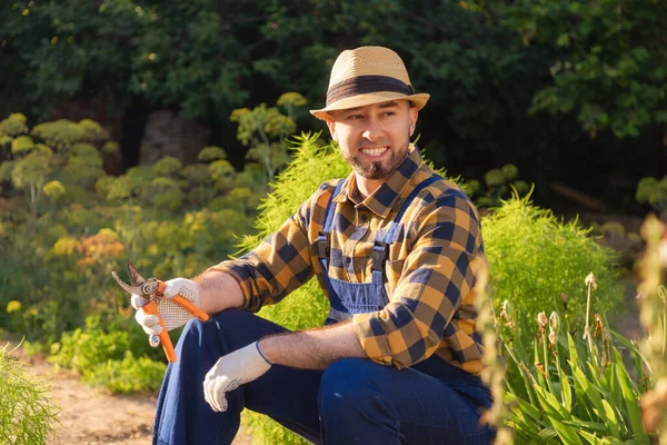 Portrait of bearded, handsome happy gardener in uniform and a straw hat sits and relax, holding a pruner. In the background is a backyard and a kitchen-garden. The concept of gardening.