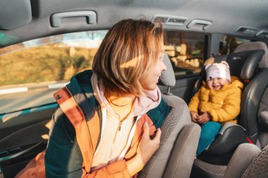 Smiling mother looking back to her cute happy little child is sitting at safe childrens backseat. The concept of safety during travel by car.