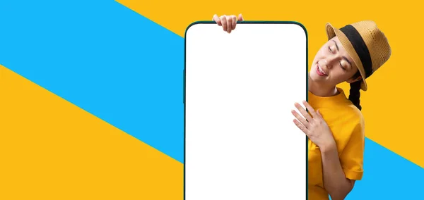 Web banner of digital advertising and social media. A young Caucasian pretty woman wearing straw hat holds a big smartphone with mock up. Isolated on yellow and blue background. Copy space. The concept of online shopping and website template