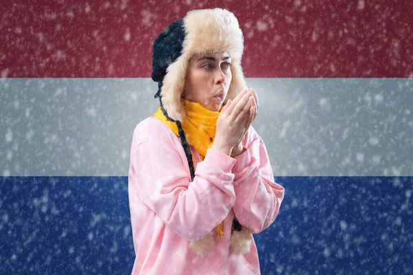 Energy crisis in Europe. Woman in hat and scarf is warming with breathing. In the background is the flag of Netherlands and a snowstorm. Concept of sanctions, refusal of gas and renewable energy.