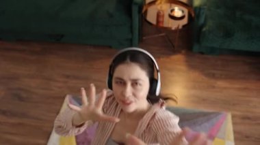 Top view of happy young Caucasian woman wearing headphones singing and dancing looking at camera. Home weekend leisure