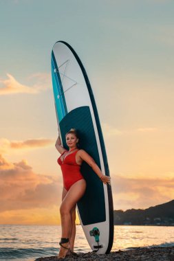 Plus sized sexy tanned woman in red bikini poses with sup board at beach. Sunset sky on the background. Vertical orientation. The concept of surfing and summer vacation.