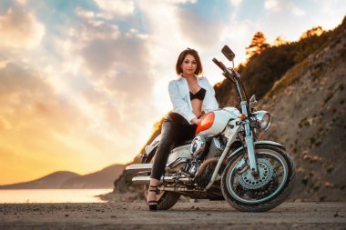 Beautiful sexy adult woman with a high heels and leather pants, posing sitting on motorcycle. Epic sunset sky on the background. The concept of Motorcyclist Day and moto travel.