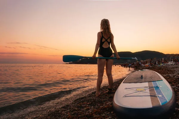 Young woman holds a paddle of sup board at the beach. Back view. Copy space. Sunset at the background. The concept of summertime, sport and vacation.