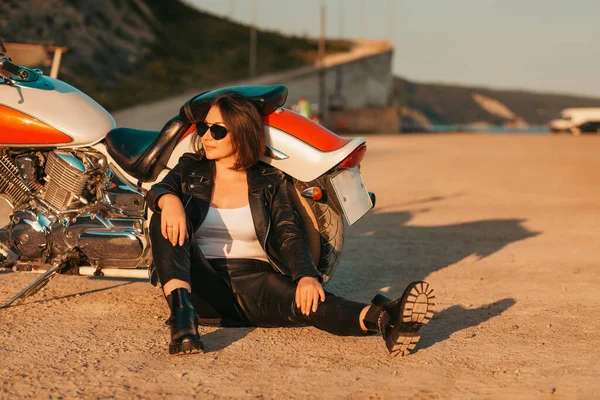 Pretty confident woman in a leather motorcycle outfit sitting on the ground near a motorcycle. The concept of the Motorcyclist Day and feminism.