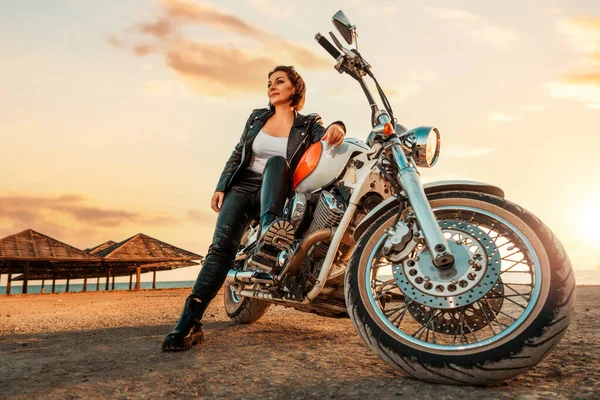 A beautiful independent woman in a leather clothes sitting on a motorcycle. Bottom view. Epic sunset on beach at the background. The concept of motorcycle trip and feminism.