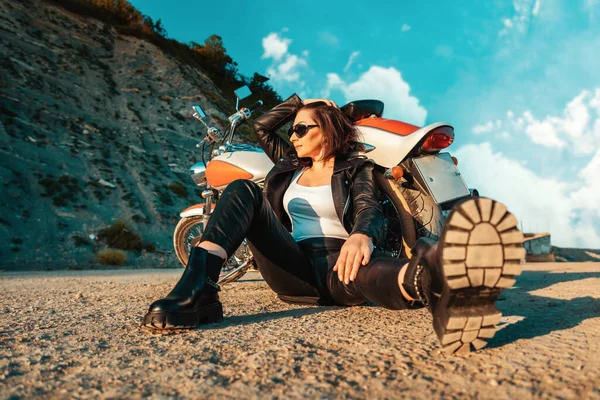 A cool independent woman in a leather motorcycle outfit poses sitting on the ground near a motorcycle. Low angle view. The concept of the motorcycle travel and feminism.