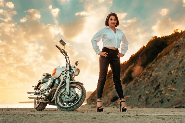 Self confident beautiful woman in high heels and leather pants posing at background with motorcycle and epic sunset sky. The concept of Motorcyclist Day.