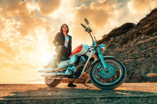 A beautiful confident adult woman in a leather clothes sitting on a vintage motorcycle. Bottom view. Epic sunset on the background. The concept of motorcycle travel and feminism.