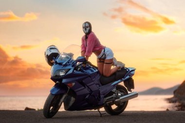 Sexy woman in a balaclava with skull poses on a tourist motorcycle. Beautiful golden sunset in the background. The concept of moto travel and motorcycle world day.