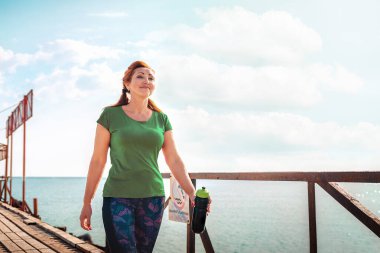 Spor kıyafetli yetişkin bir kadın, elinde bir sallayıcıyla rıhtımda yürüyor. Uzayı kopyala Açık hava yürüyüşü.