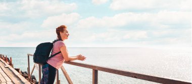 Pankart. İskelede sırt çantasıyla poz veren spor kıyafetli mutlu bir yetişkin kadın. Arka planda, deniz ve gökyüzü. Uzayı kopyala Aktif yaşam tarzı ve özgürlük kavramı.