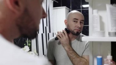 Skin care. Portrait of handsome young caucasian man shaving face with electric shaver, looking at himself in mirror and smiling. Beauty routine.