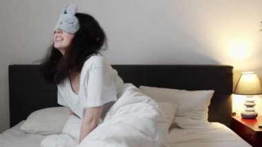 Awaked angry young Caucasian woman in sleep mask covers her ears with pillow, shakes fists and screams because of neighbours making noise and interfering with sleep. Concept of insomnia and sleepless.