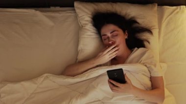 Uyku vakti. Beyaz tenli genç bir kadının yatakta uzanmış akıllı telefon kullanışının en üst görüntüsü. Uykusuzluk ve sosyal medya bağımlılığı kavramı.